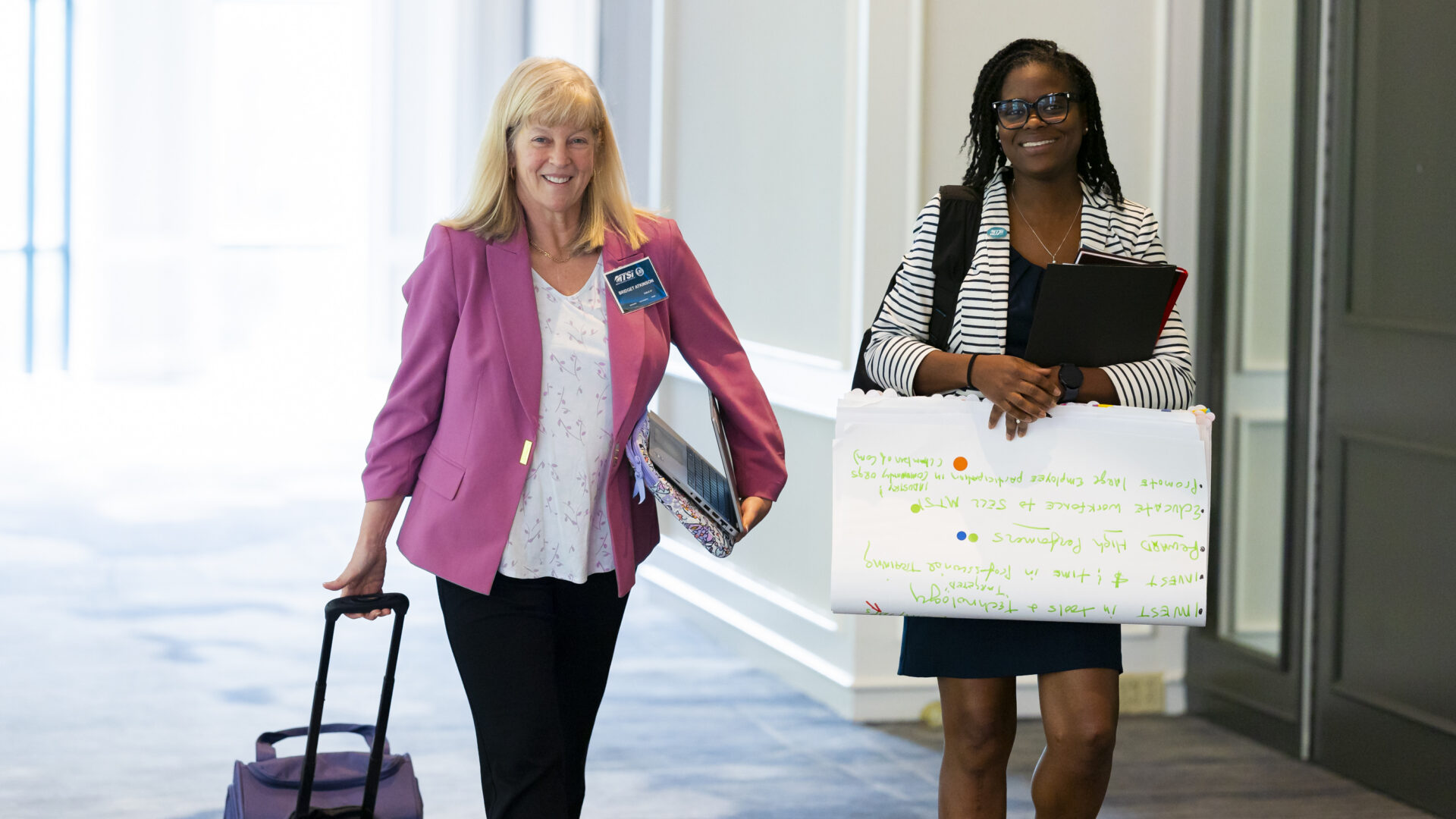 Two professionals walking through conference venue carrying presentation and meeting materials.
