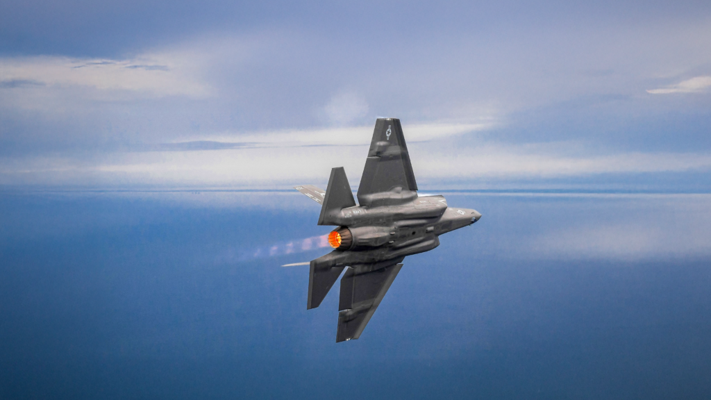 F-35 fighter aircraft flying above the ocean with afterburner engaged.