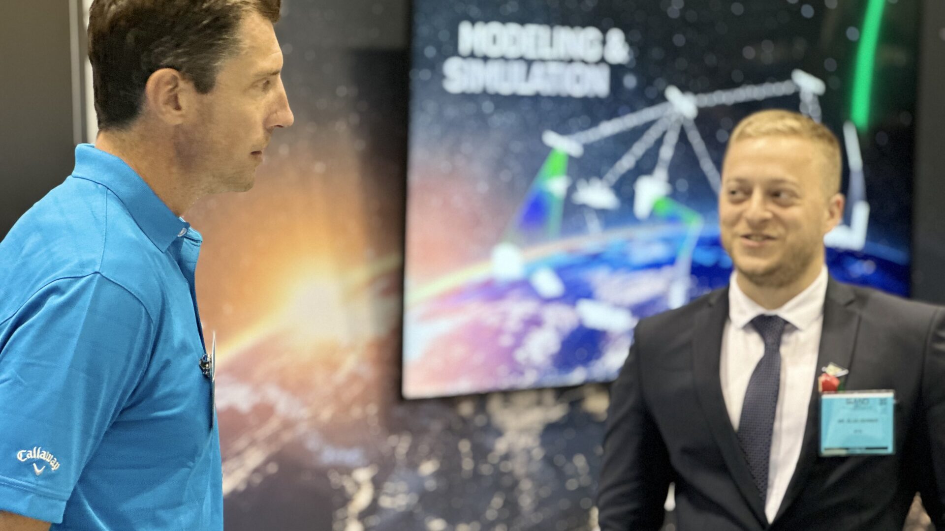 Two professionals speaking at industry booth with modeling and simulation display in background.