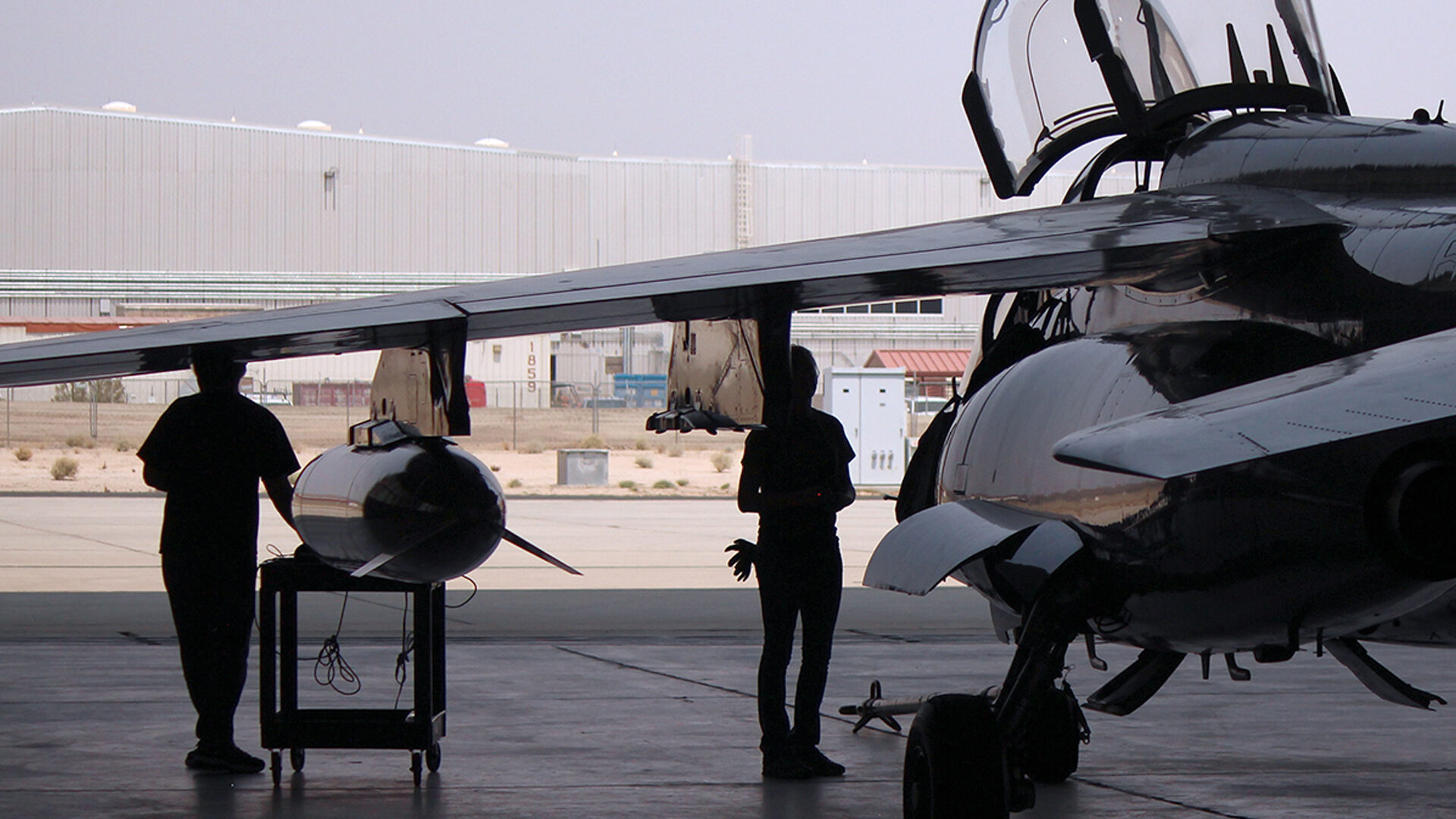 Personnel standing beside military aircraft and mounted equipment inside aircraft hangar.