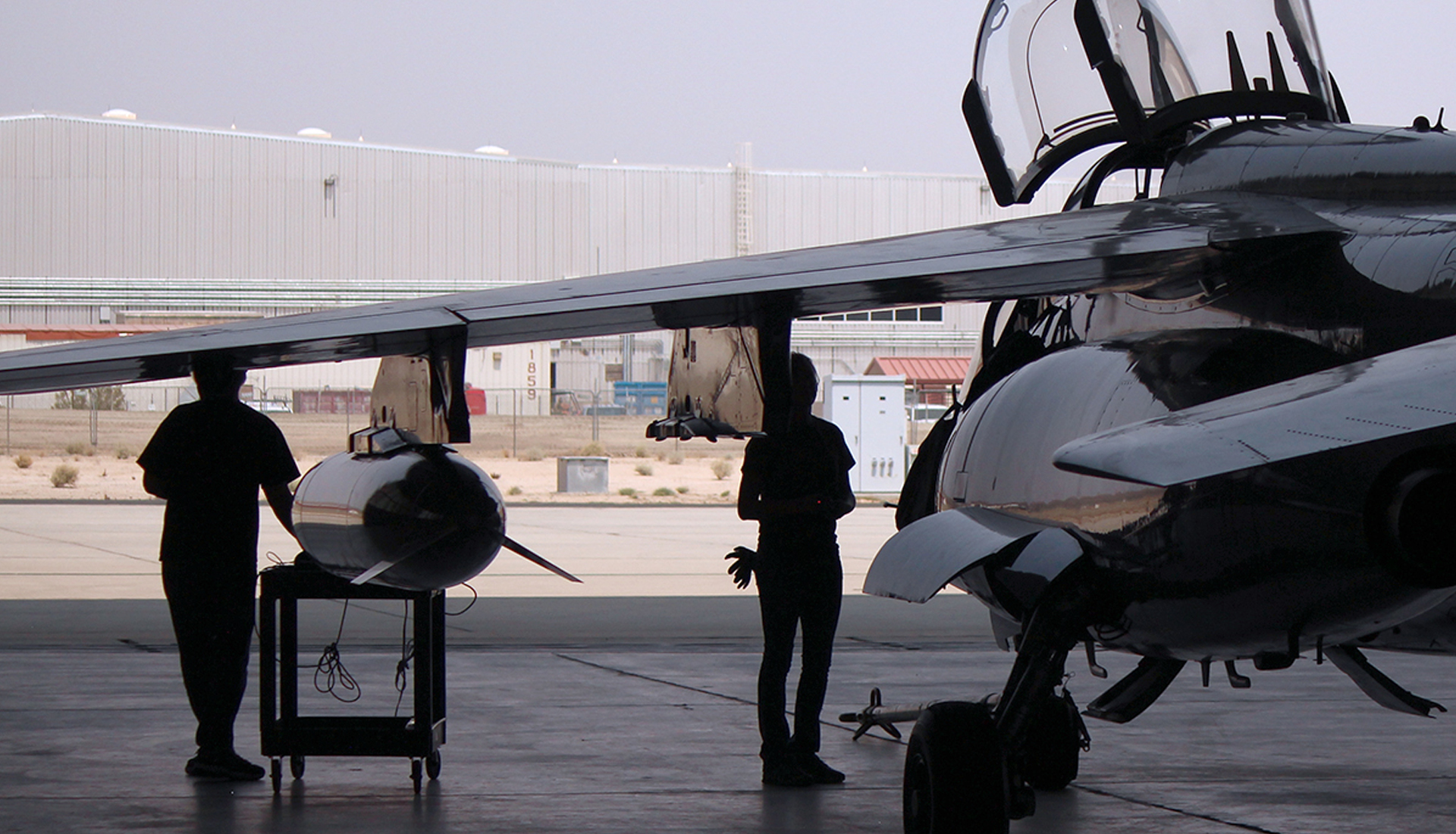 Personnel standing beside military aircraft and mounted equipment inside aircraft hangar.