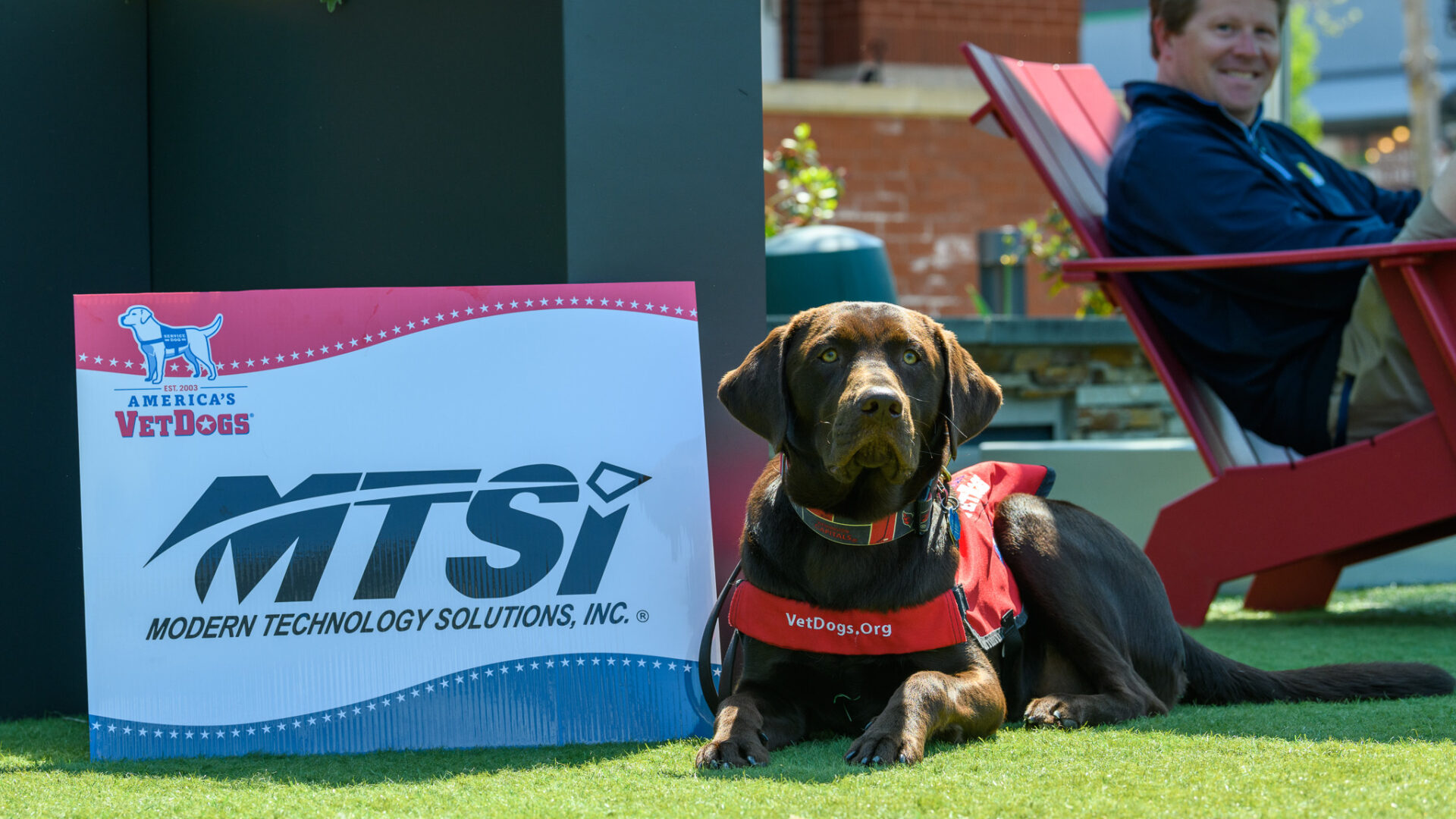 Service dog wearing vest beside company sign at outdoor community event.