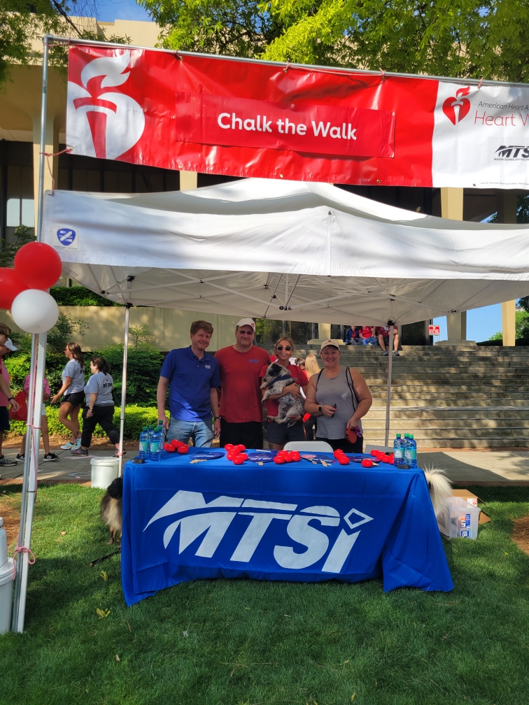 Community outreach booth at outdoor charity walk event with participants standing beneath event banner.