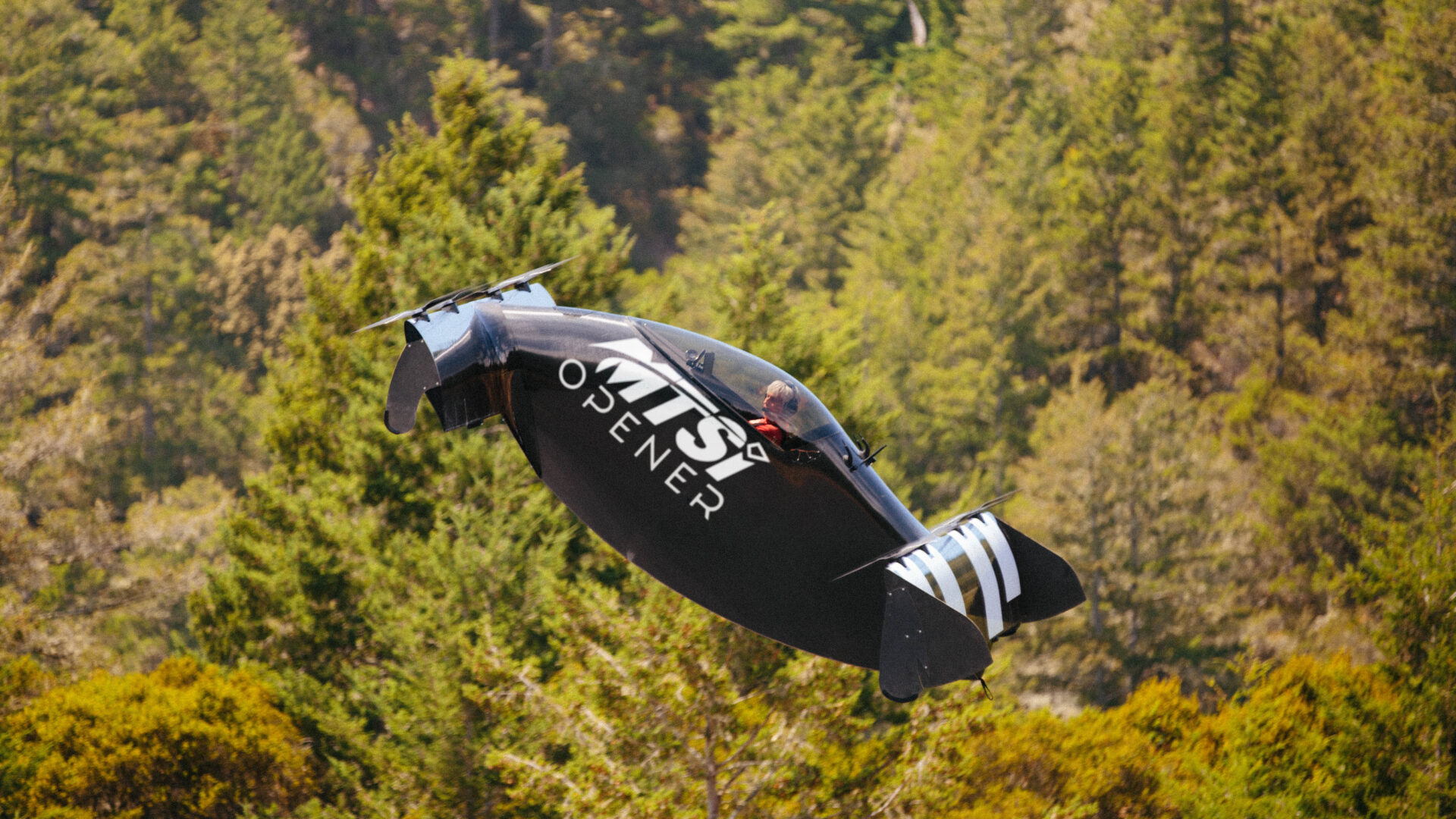 Experimental vertical takeoff aircraft flying above forested terrain.
