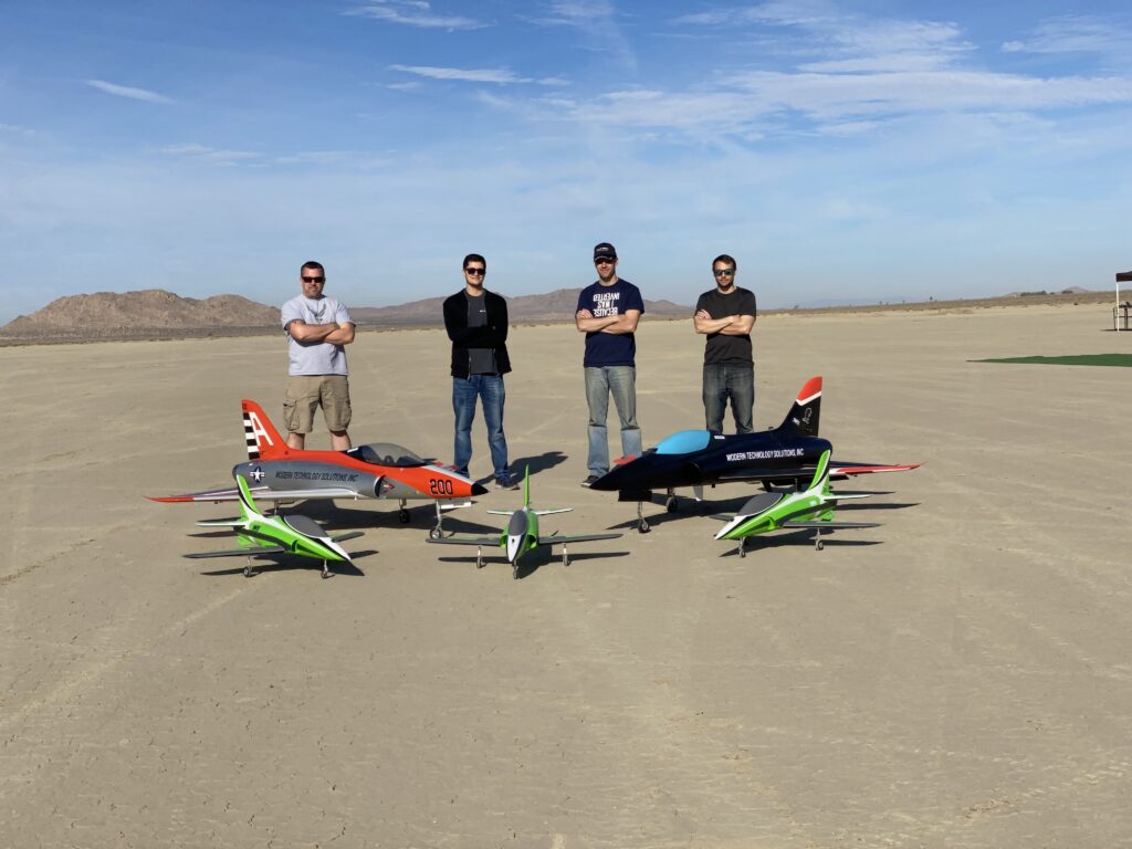 Group standing on desert airfield with experimental aircraft and unmanned aerial vehicles.