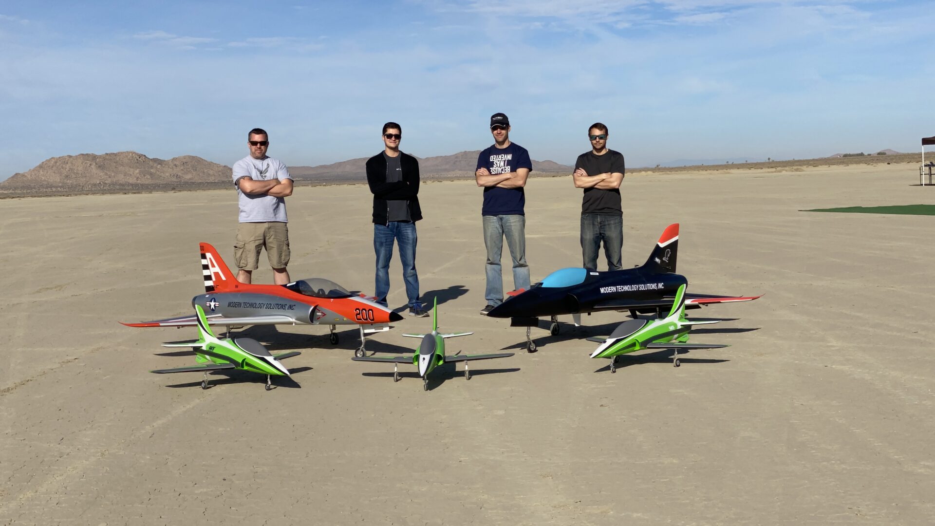 Group standing on desert airfield with experimental aircraft and unmanned aerial vehicles.